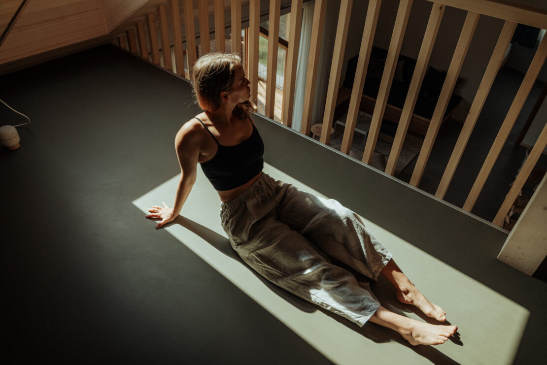 In the gallery under the vaulted ceiling, a woman sits with her legs stretched out and rests her hands on the floor behind her
