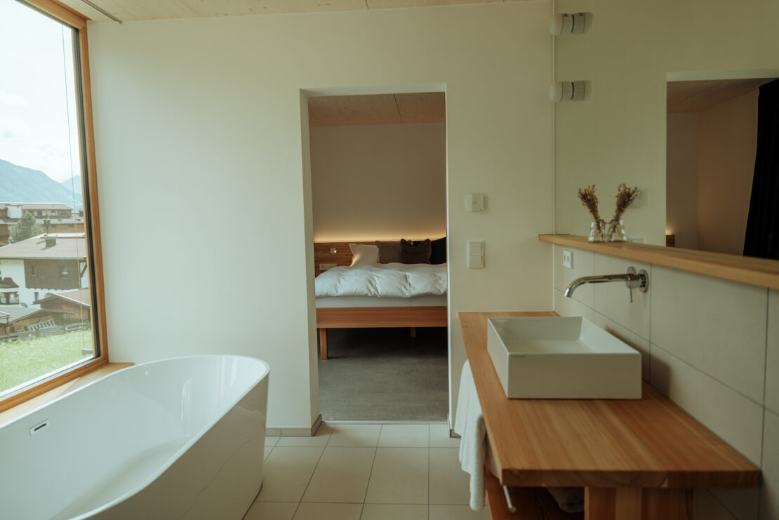 Bathroom with washbasin on larch wood, large mirror and panoramic window, in front of it a bathtub, in the background the bedroom with double bed
