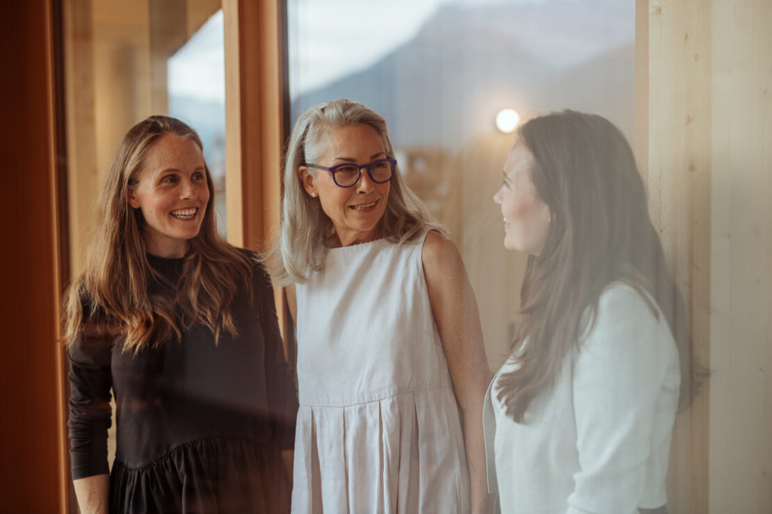 Three women with long hair stand in front of a glass front and smile at each other in a friendly manner.