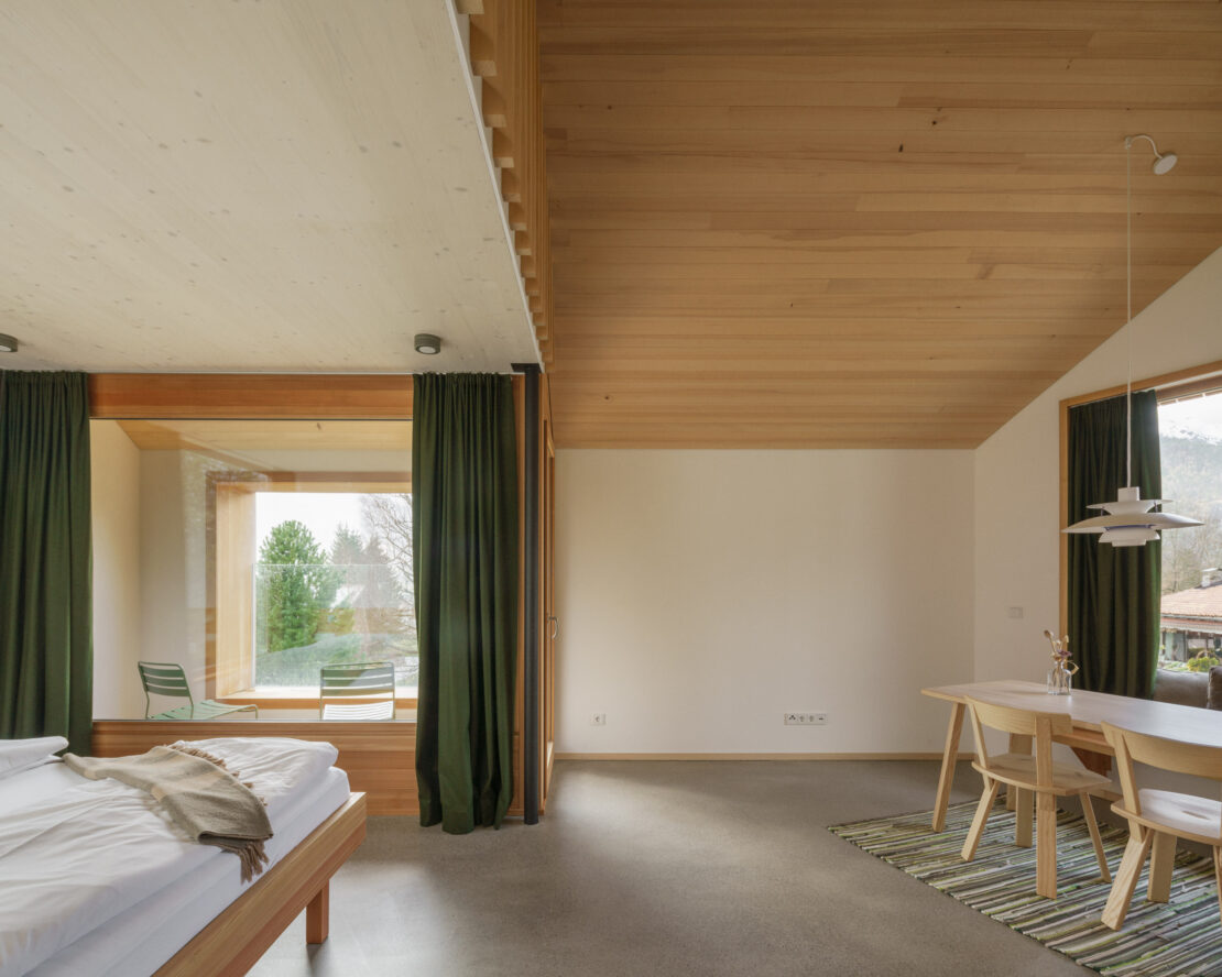 A living/sleeping area with a large panoramic window with green loden curtains and a double bed made of solid wood, the grey floor is polished concrete