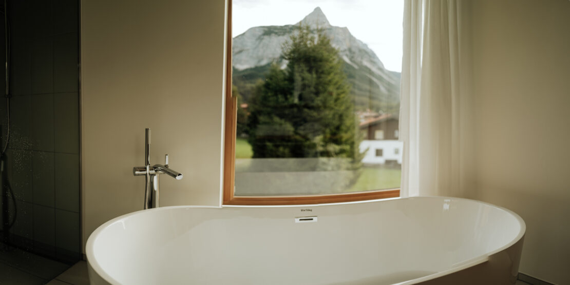 Free-standing bathtub in front of a deep window front with a view of conifers and the Ehrwald mountain landscape, Haus Aerli Ehrwald