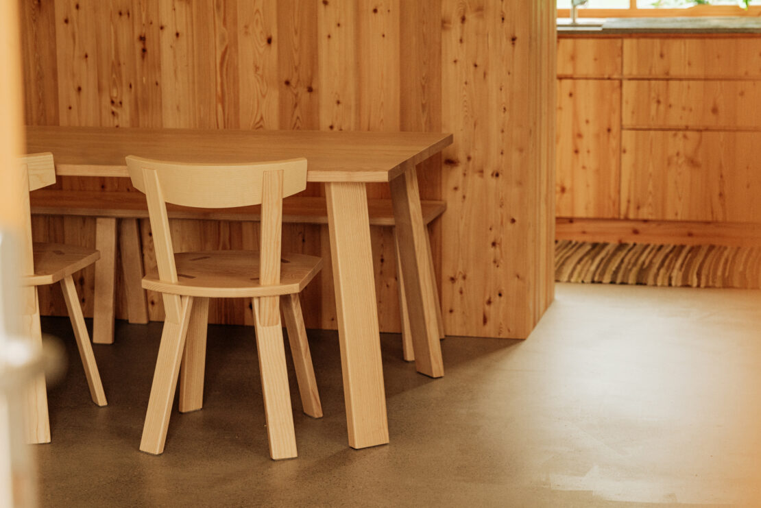 House Aerli Detail view of a wooden table with wooden chairs and a kitchen front made of larch wood, a pile carpet lies on the screed floor