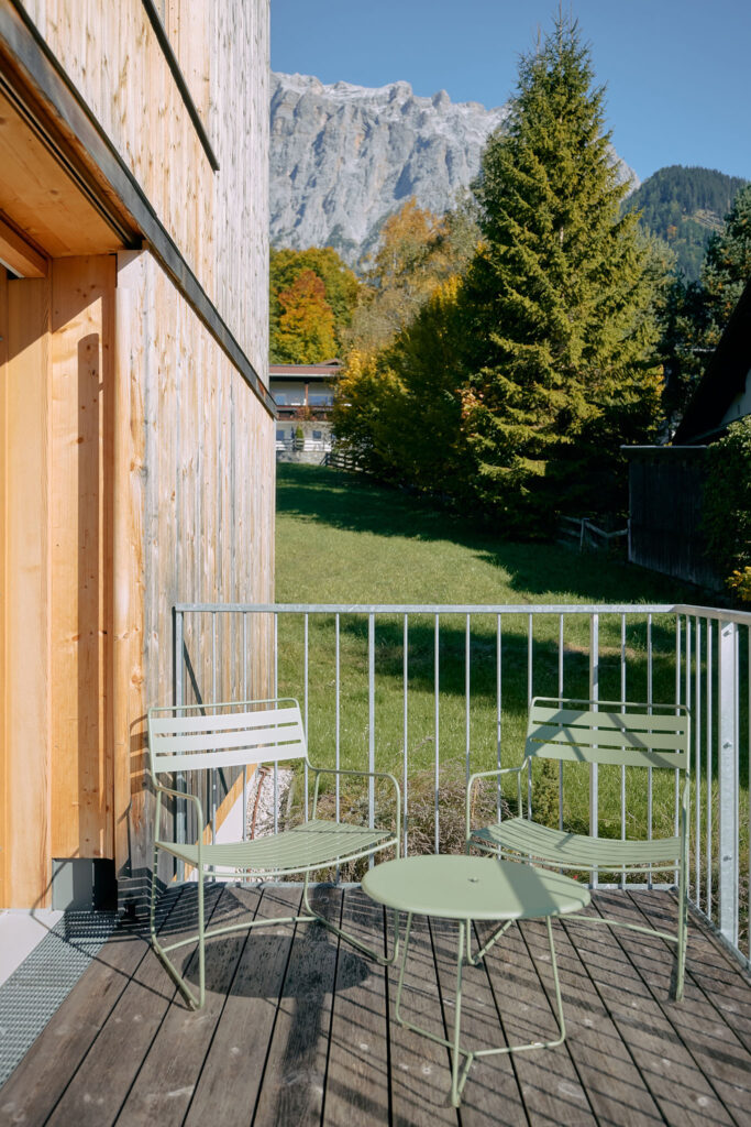 At design holiday home Aerli two light-green metal bistro chairs and a small table sit on a sunny wooden balcony, overlooking a grassy slope, evergreen trees, and a towering, rocky mountain peak.