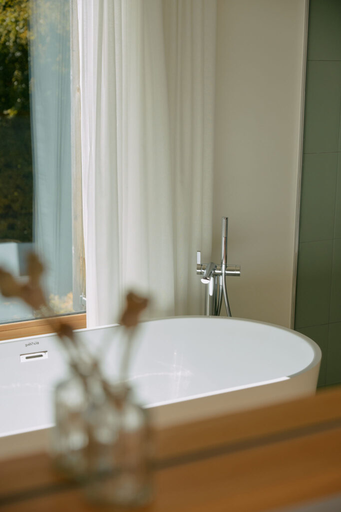 Detail of a freestanding white bathtub in front of a large window, with a vase in the foreground.
