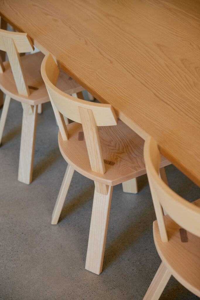 Here is the description in English: A row of light-colored, modern wooden chairs with curved backrests is tucked neatly under a long, matching wooden table on a smooth gray floor.