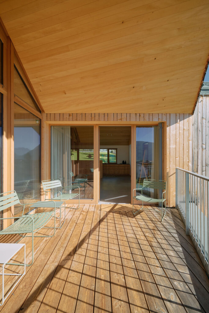 A spacious wooden balcony at holiday house Aerli, covered by a matching wooden ceiling, is furnished with light-green metal chairs and opens into a bright room through a large sliding glass door.