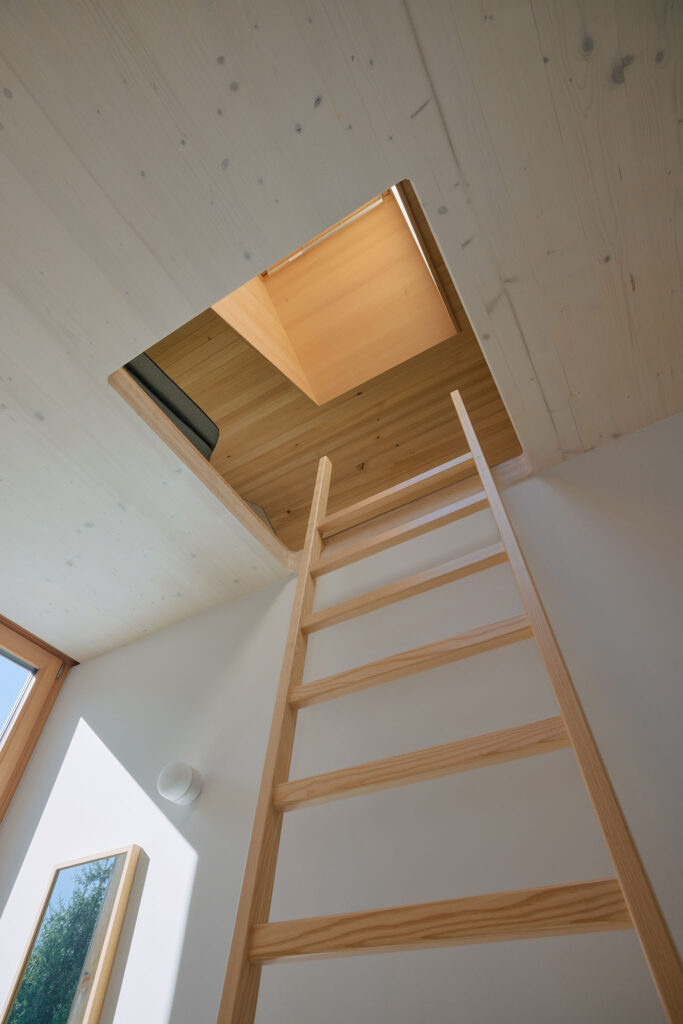 Eine Holzleiter ragt entlang einer Wand hoch unter eine Ebene unter dem Sichtdachstuhl aus Holz.