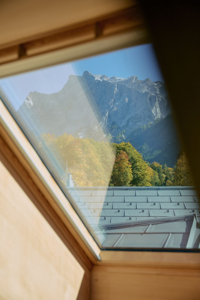 View from the wooden-framed skylight of Haus Aerli onto the Zugspitze mountain range with autumnal forest in the foreground