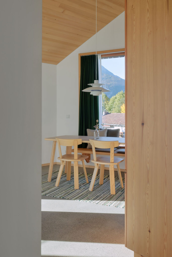 View from a distance of the dining area with carpenter-made furniture and lamp by Louis Poulsen under exposed roof trusses, behind it a window front with loden curtain