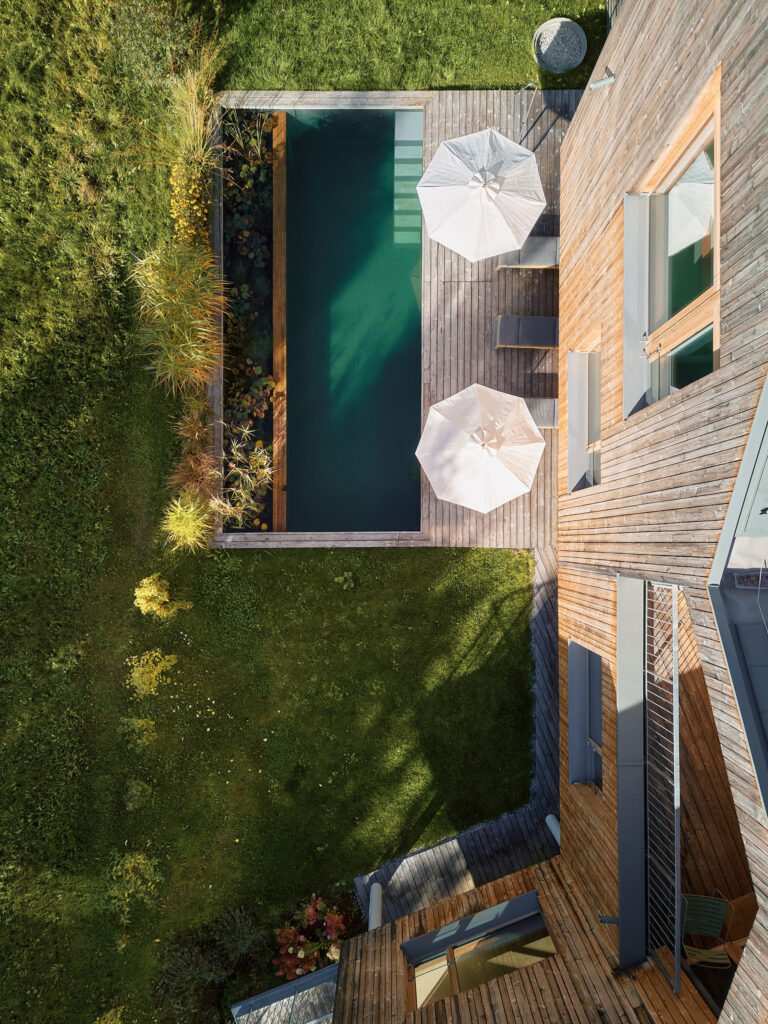 Blick von oben auf das Design-Ferienhaus Aerli mit Fichtenfassade, Garten mit Sträuchern und Naturpool mit begrünter Regenerationszone, Sonnenterasse mit weißen, runden Sonnenschirmen und Sonnenliegen