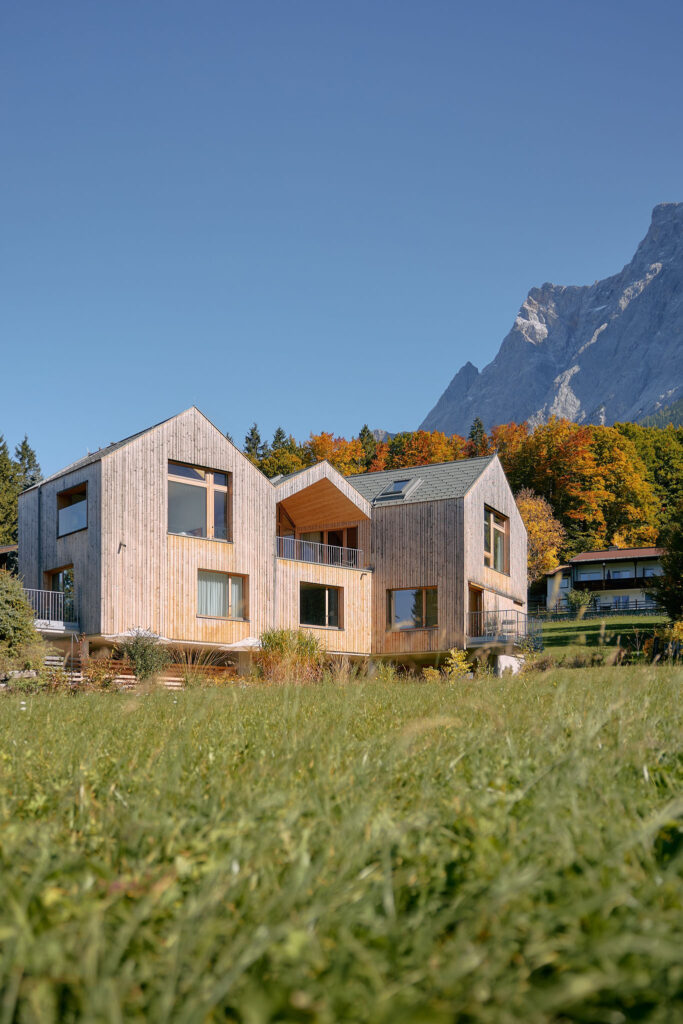 Holiday house Aerli, with its wooden façade and three gabled roofs, stands in front of an autumnal forest and the Zugspitze massif.