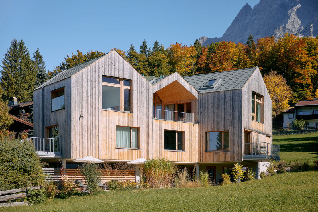 Haus Aerli mit Holzfassade und drei Satteldächern steht vor herbstlichem Wald und Zugspitzmassiv