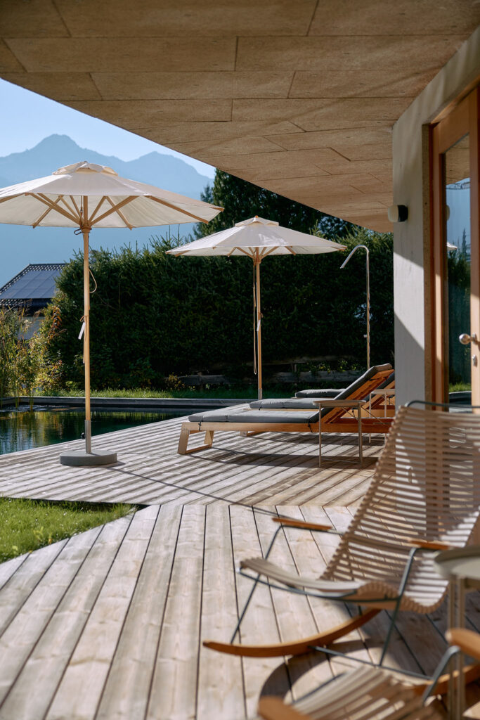 Wooden sun terrace with loungers, rocking chairs and white parasols against a mountain backdrop.
