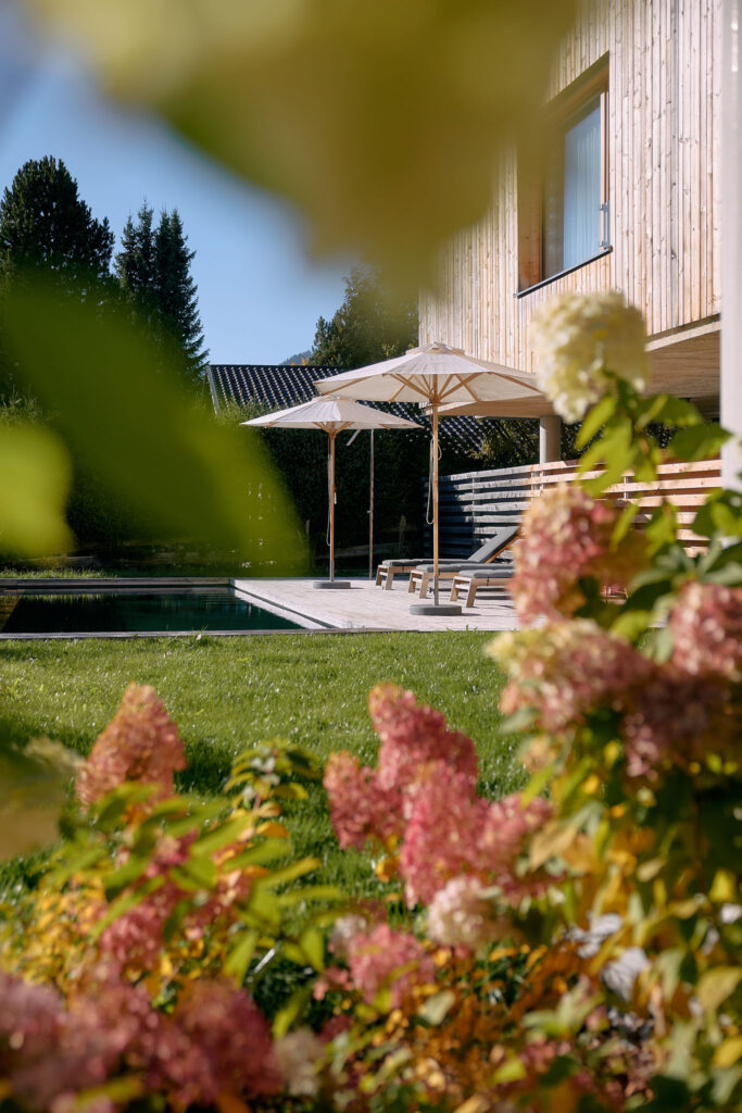 Seen past blurry pink flowers in the foreground, a sunlit garden features a wooden patio with two large umbrellas and lounge chairs next to a green lawn and a modern timber-clad building.