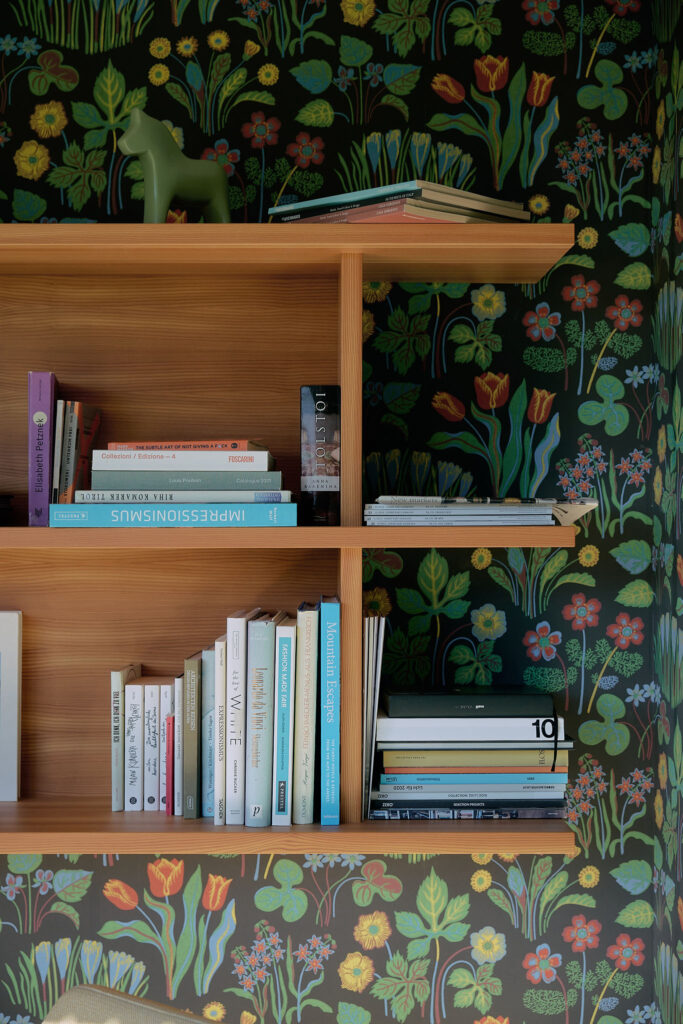 A larch wood bookshelf with multiple compartments holds stacks of books, set against a dark wallpaper featuring a vibrant, colorful floral and plant pattern designed by Josef Frank.