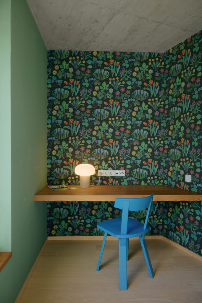 Blauer Holzstuhl von der Tischlerei Hussl, Schreibtisch aus Lärchenholz, darauf eine Designlampe, rundum florale Tapete des Designers Josef Frank.