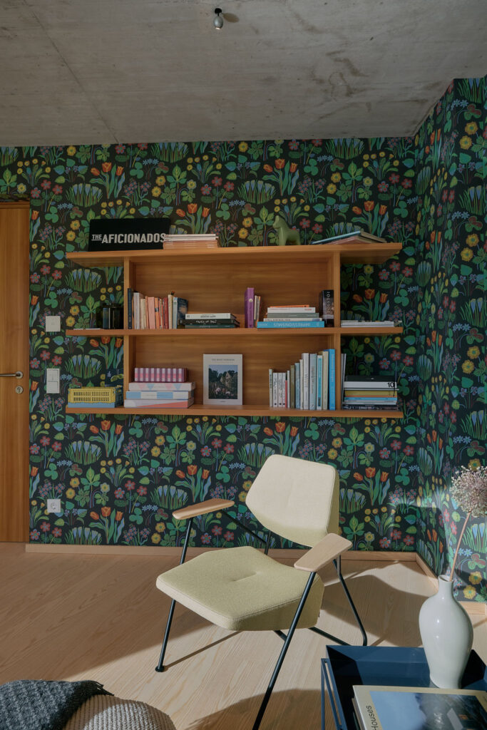 At design holiday house Aerli a modern, light-colored chair sits in a patch of sunlight on a wooden floor, positioned in front of a wall covered in dark, colorful plant-patterned wallpaper by Josef Frank that also features a wooden shelf holding books.