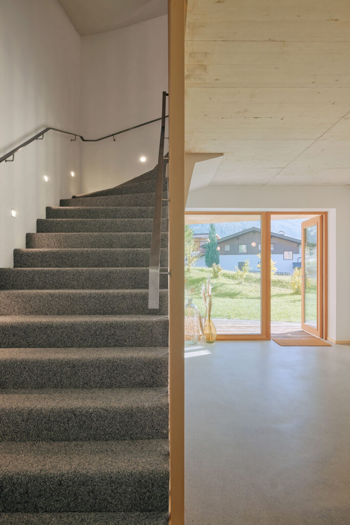 On the left, stairs with grey carpet and floor lamps; on the right, screed floor and view through the floor-to-ceiling window front into the garden of the Aerli designer holiday home.