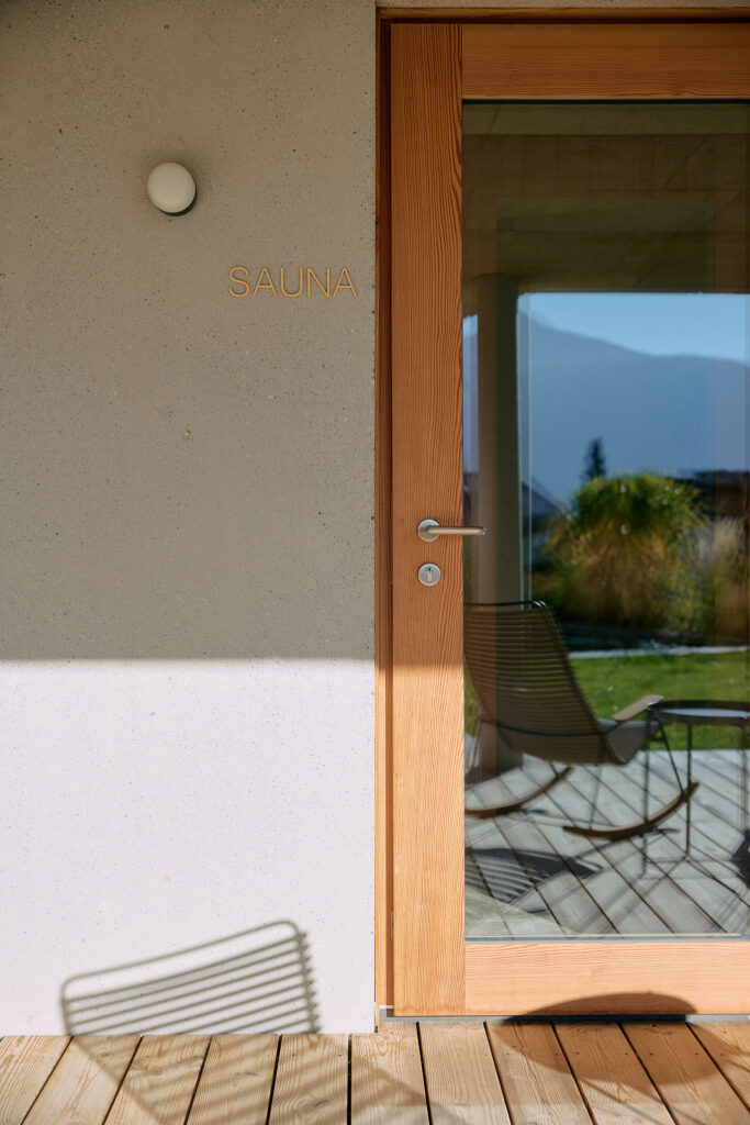 The entrance to a modern sauna features a wooden-framed glass door next to a light-grey wall with the word 